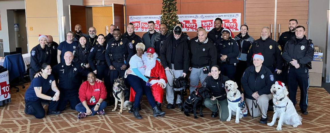 Large group of TUPD officers and dogs posing in front of a lit Christmas tree and a sign that reads "Toys for Tots".