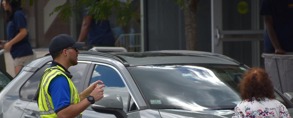 A security officer in a high-visibility vest directs a gray car