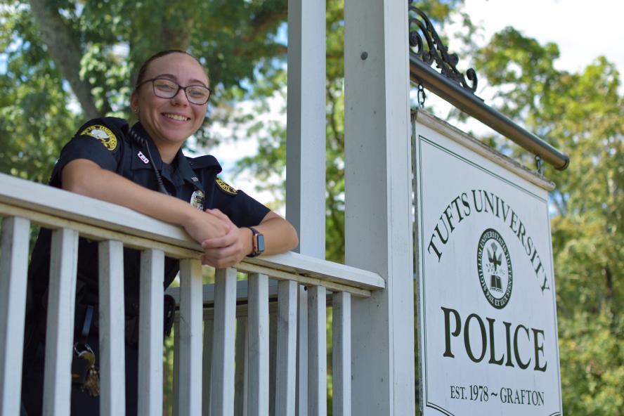 A female TUPD officer wearing glasses is standing on the porch next to a sign that reads "Tuft's University Police"
